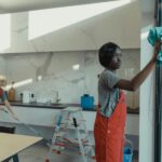 Two women cleaning a modern kitchen, wiping glass surfaces and mopping the floor.